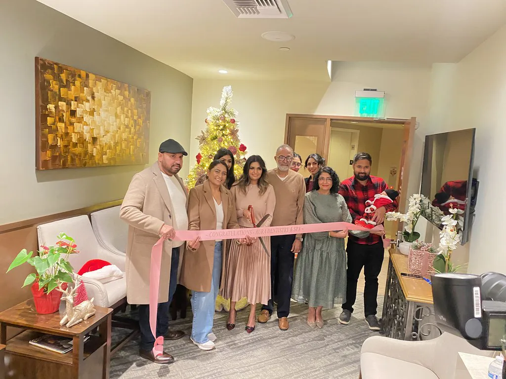 A group of eight people stand in a decorated room holding a pink ribbon. A Christmas tree and holiday decorations are visible. The ribbon reads 