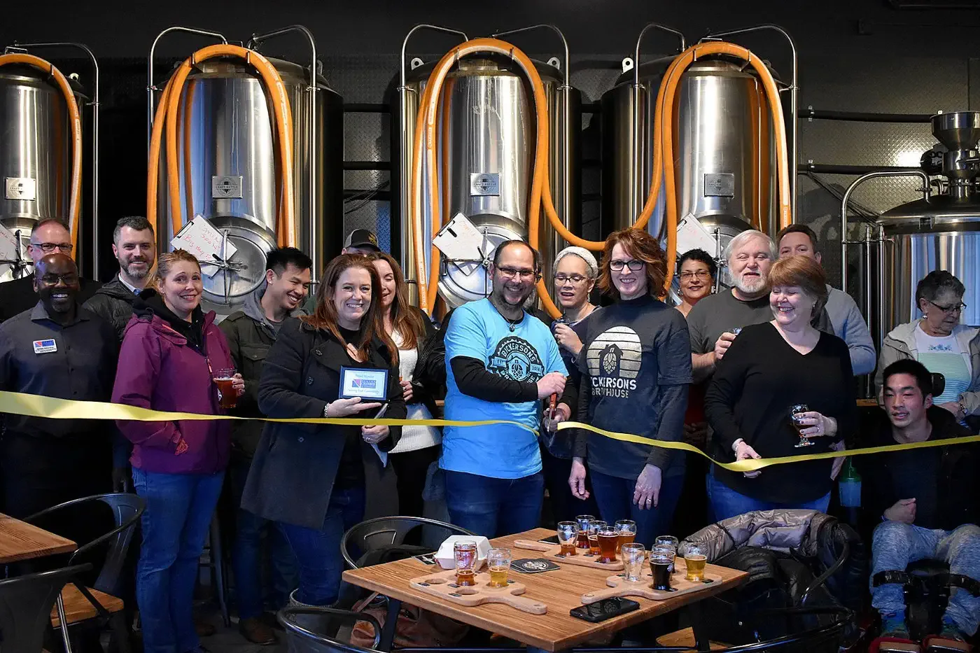 A diverse group of people smiling and celebrating inside a brewery, with large fermentation tanks behind them. A person in the center holds scissors, cutting a yellow ribbon.