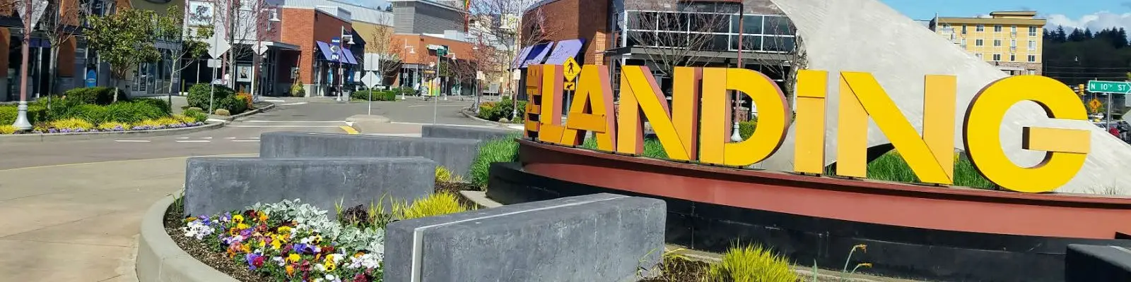 Large yellow letters display LANDING, surrounded by modern buildings and vibrant flowers. The street curves ahead, lined with trees and shops under a clear blue sky.