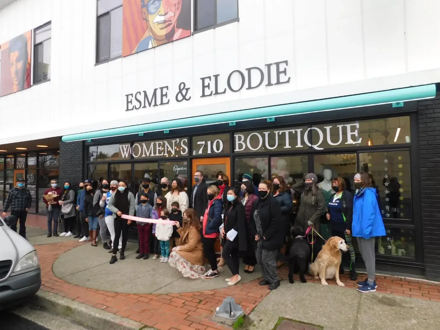 A group of people gathers for a ribbon-cutting ceremony outside a store named Esme & Elodie Women's Boutique. Most are wearing masks and two dogs are present.
