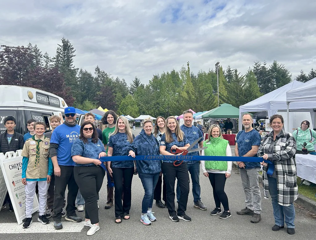 A group of people stands outdoors in front of market stalls, holding a blue ribbon for a ceremonial cutting. Trees and a coffee vehicle are in the background under a cloudy sky.