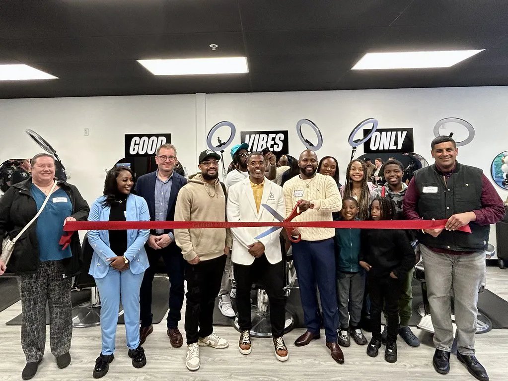 A group of people smiling and posing at a ribbon-cutting ceremony inside a salon. A man in the center holds large scissors cutting the red ribbon. Signs read 
