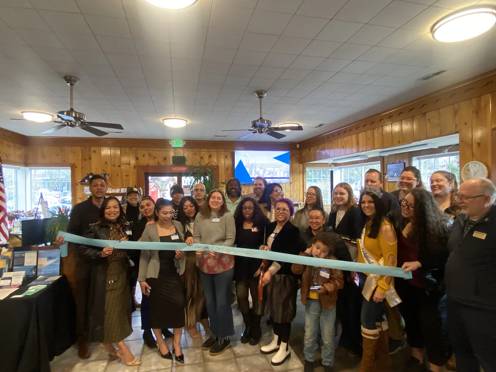 A group of people smiling and standing indoors at a ribbon-cutting ceremony. They hold a large blue ribbon in a space with wooden walls and ceiling fans in the background.