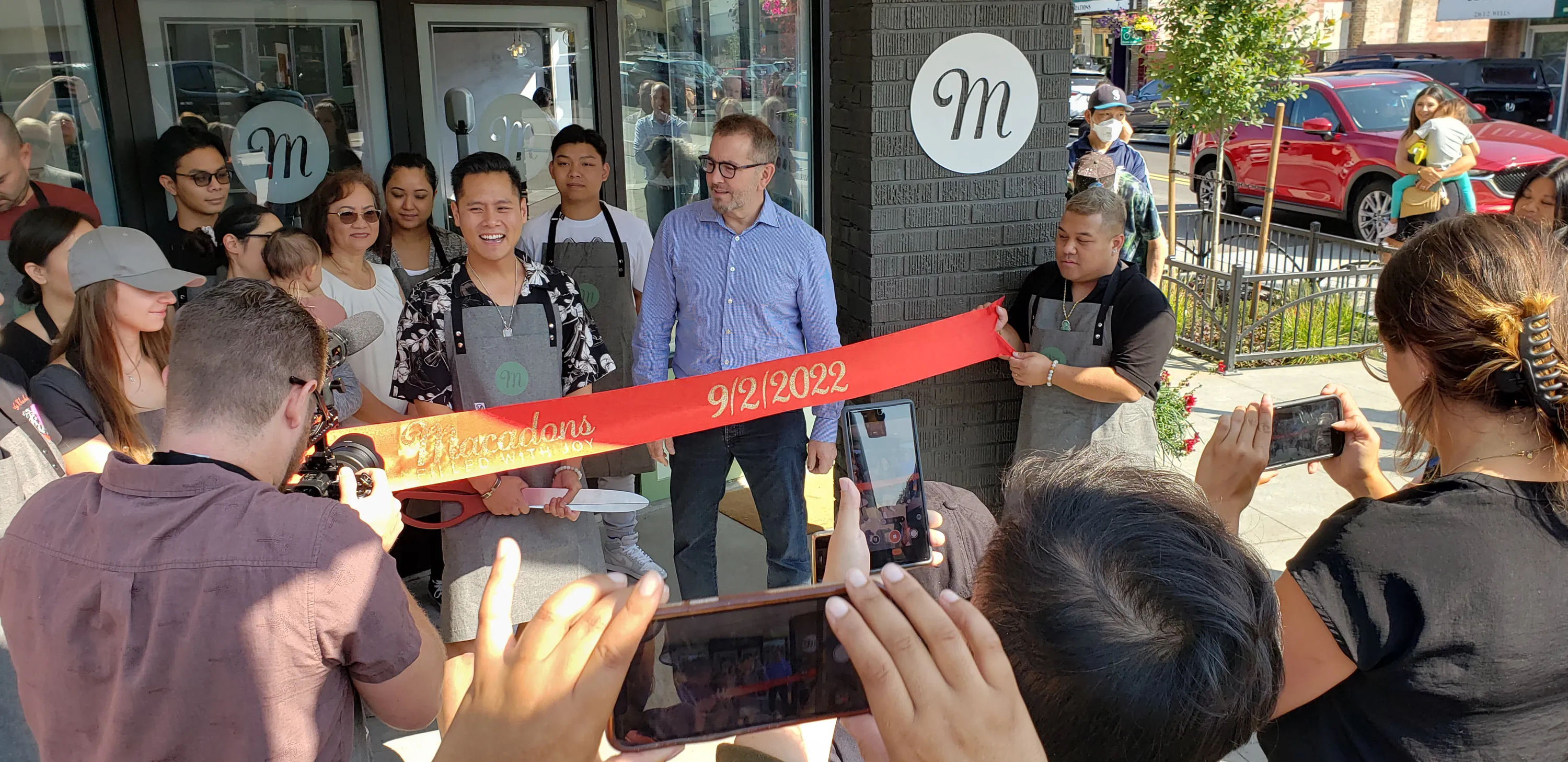 Group of people at a ribbon-cutting ceremony outside a store with a red ribbon dated 9/2/2022. A person is about to cut the ribbon while others take photos.
