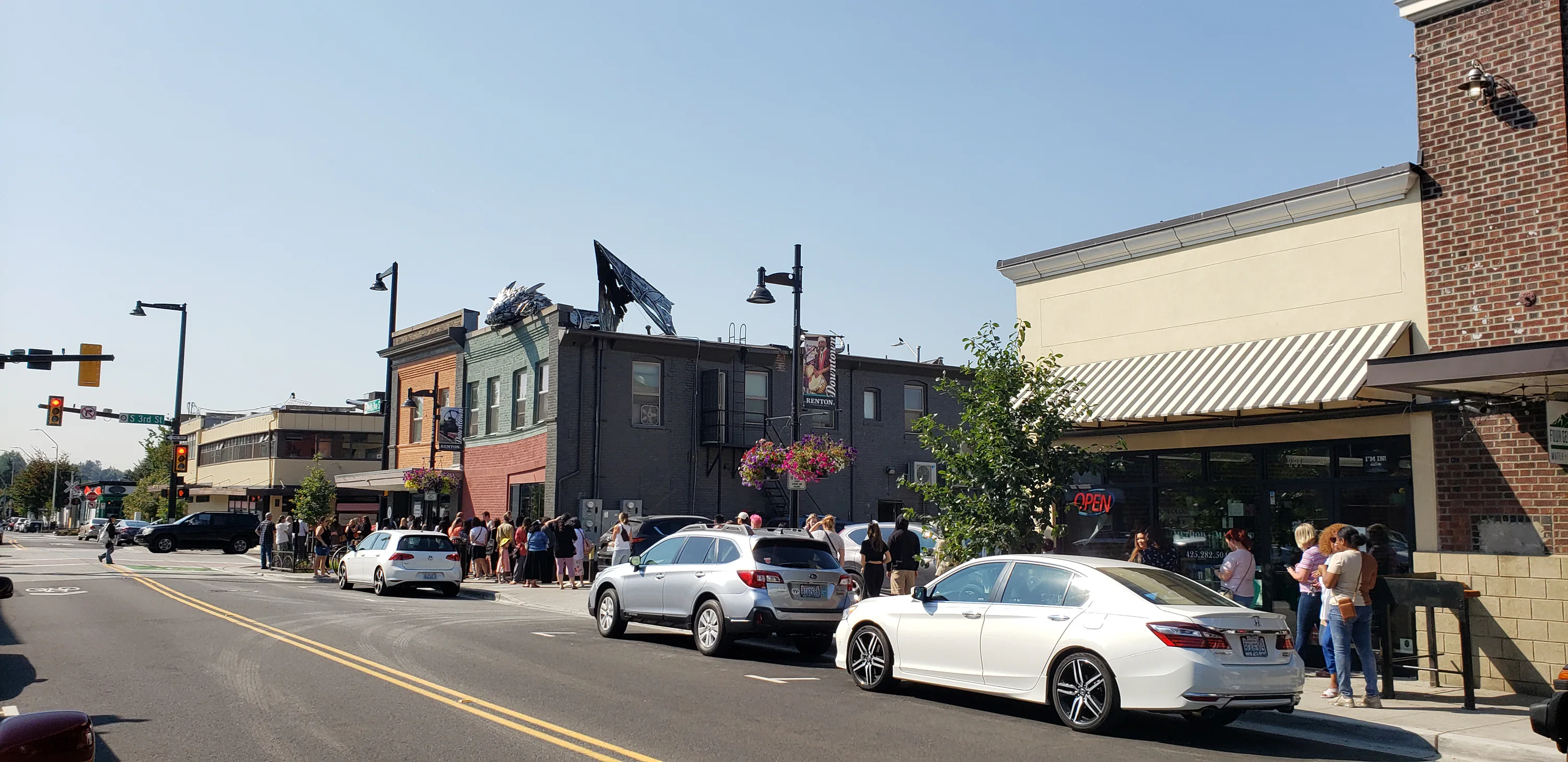 A busy street scene with cars parked along the road and a line of people in front of a brick building. A dragon sculpture decorates the roof of a colorful two-story building.
