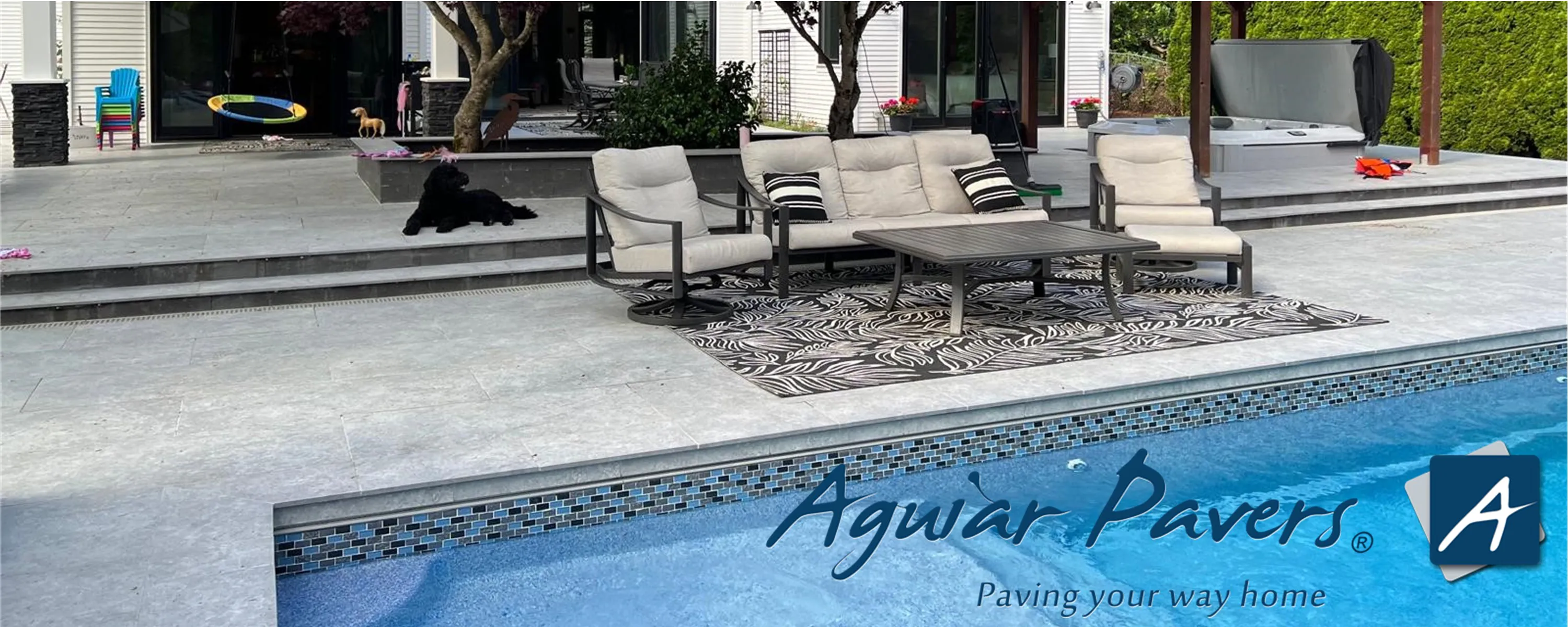 Backyard pool area with blue tiled pool, gray paver patio, beige cushioned sofa and chairs on patterned rug, coffee table, black dog on steps and a hot tub in background