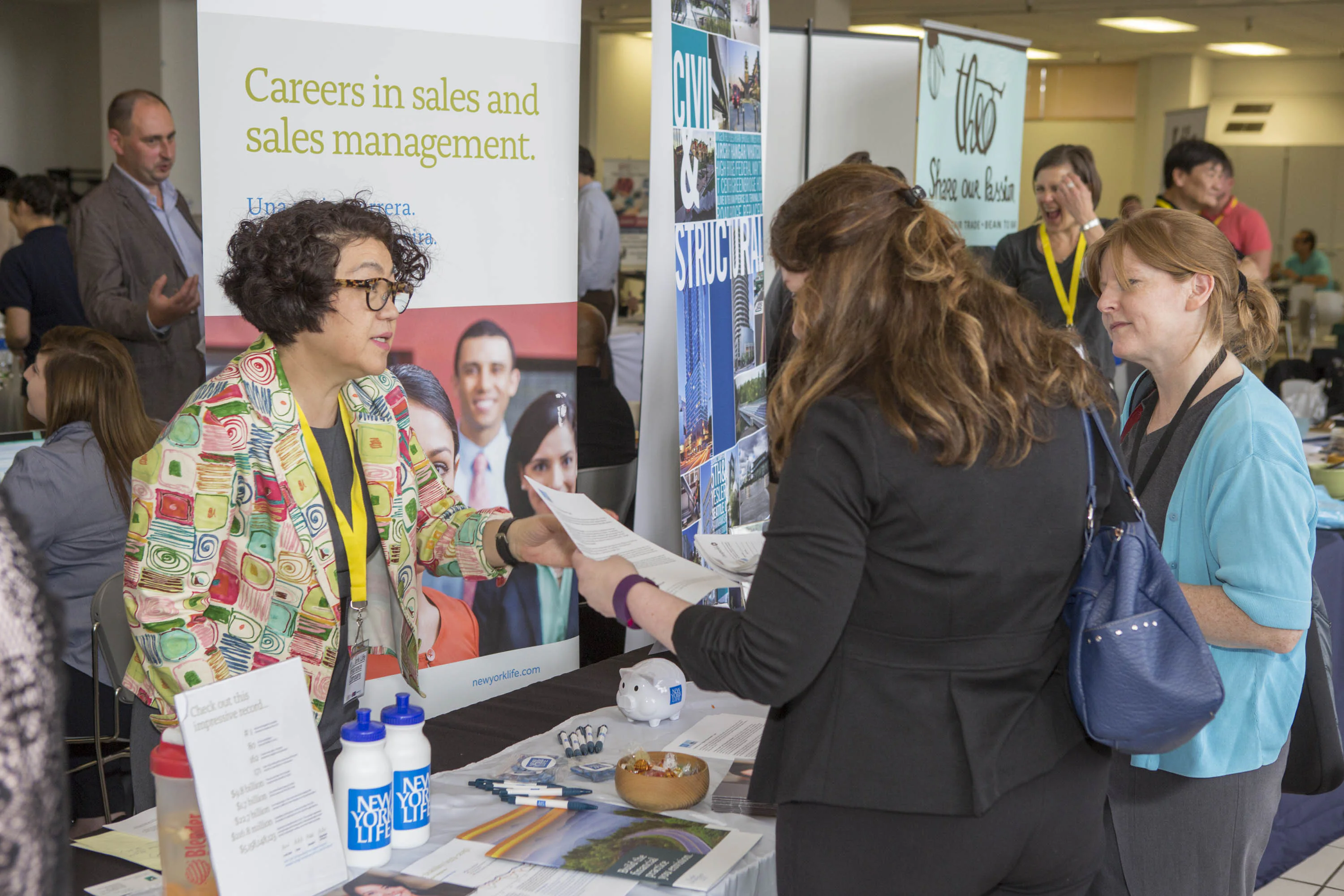 A woman in a colorful blazer talks to two attendees at a career fair booth. The booth displays materials and offers insights into careers in sales and management.