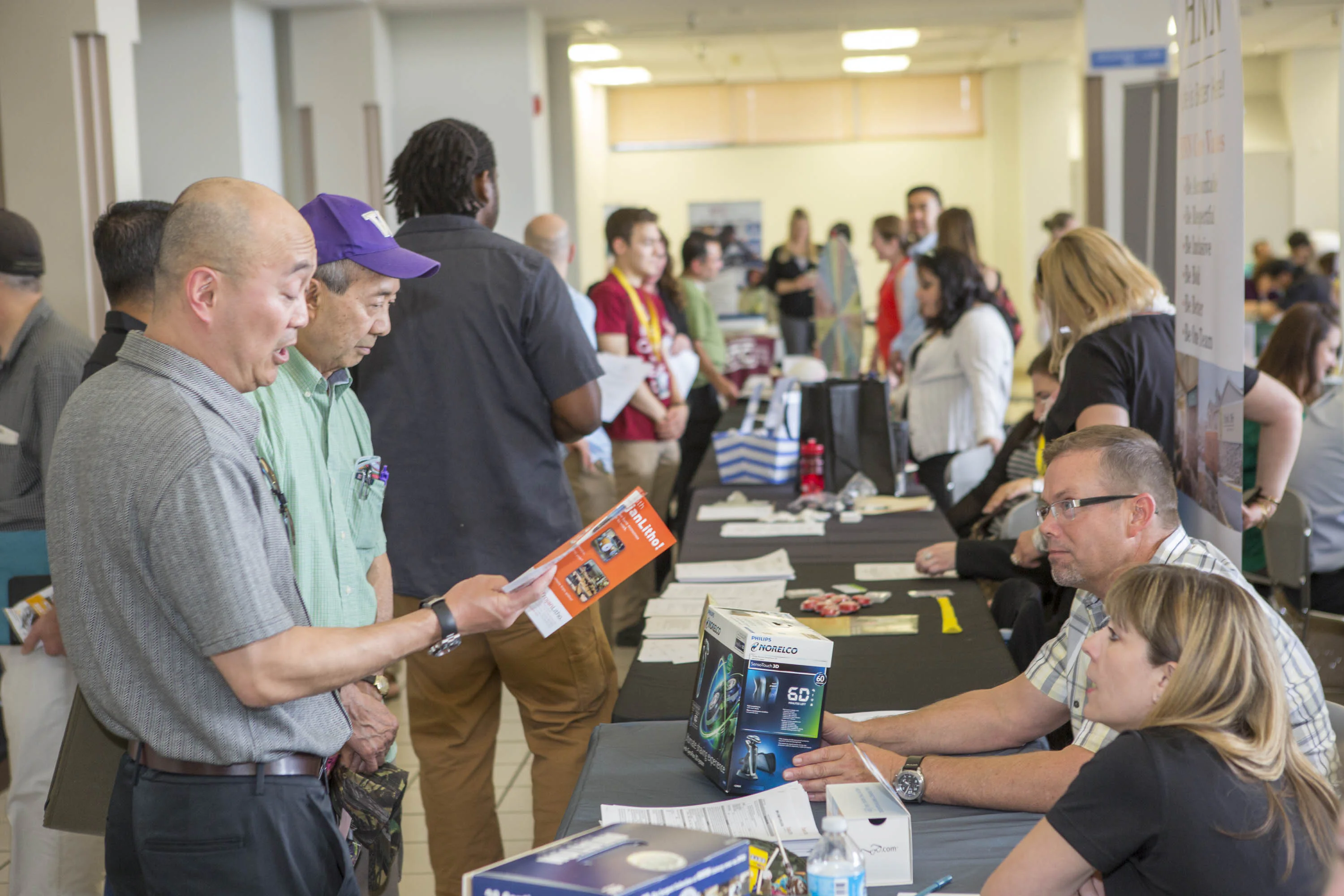 A diverse group of people participate in a bustling job fair. Attendees walk around tables where company representatives offer brochures and information about their organizations.