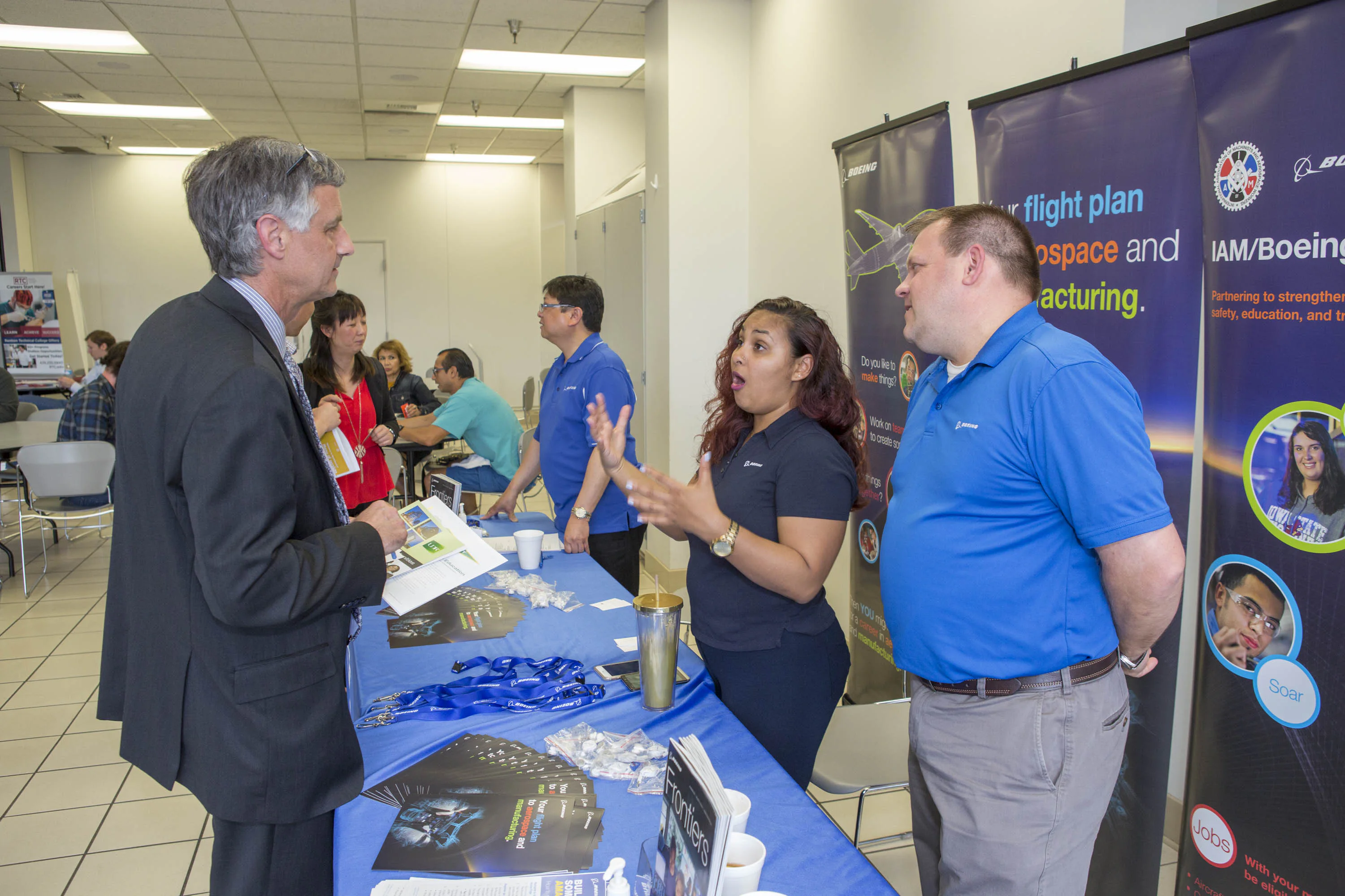 People engaged in conversation at a career fair booth. The table is covered with brochures and lanyards. Background banners display aerospace and manufacturing themes.