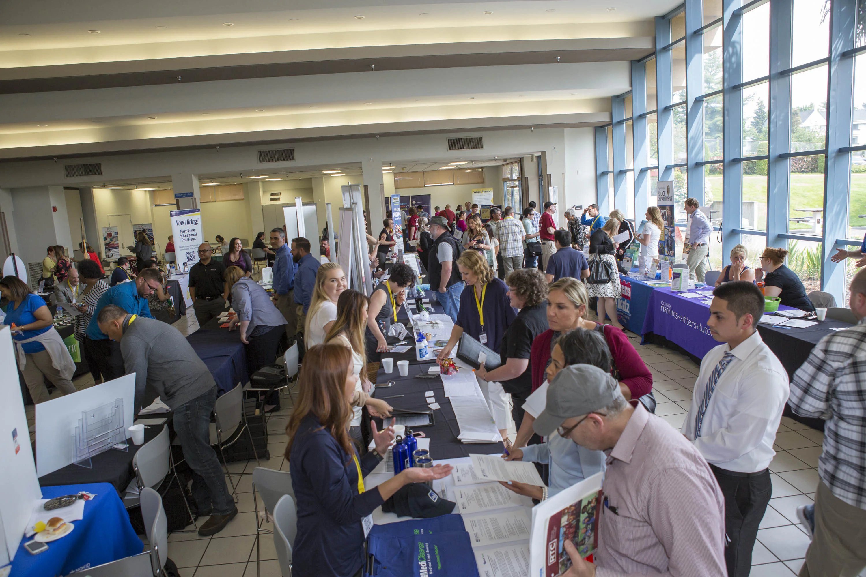 People attending a job fair in a large room with tables and displays. Attendees interact with company representatives, reviewing materials and discussing opportunities.