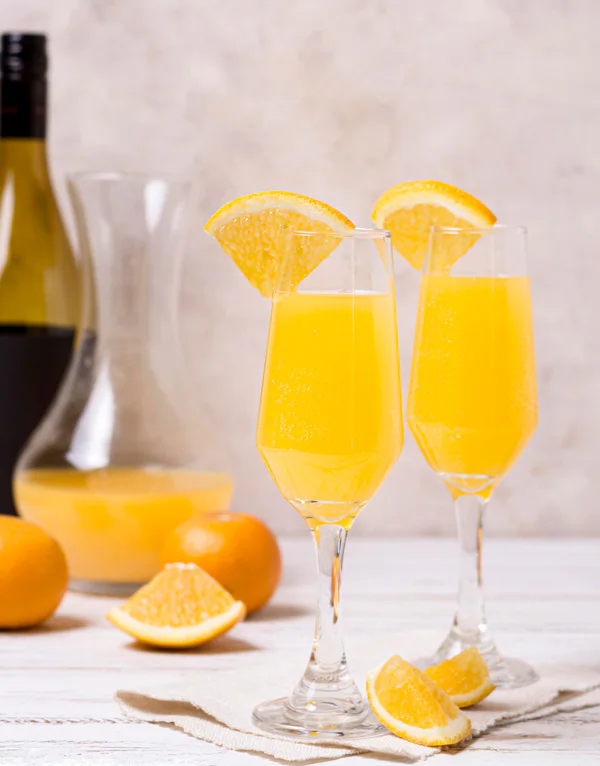Two glasses of mimosa garnished with orange slices on a white table. A bottle, carafe of juice, and whole oranges are in the background.