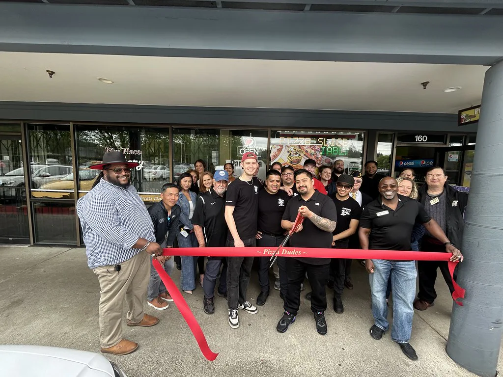 Group of people celebrating a ribbon-cutting ceremony outside Pizza Dudes storefront. A man in the center holds scissors, while others smile and gesture towards the camera.