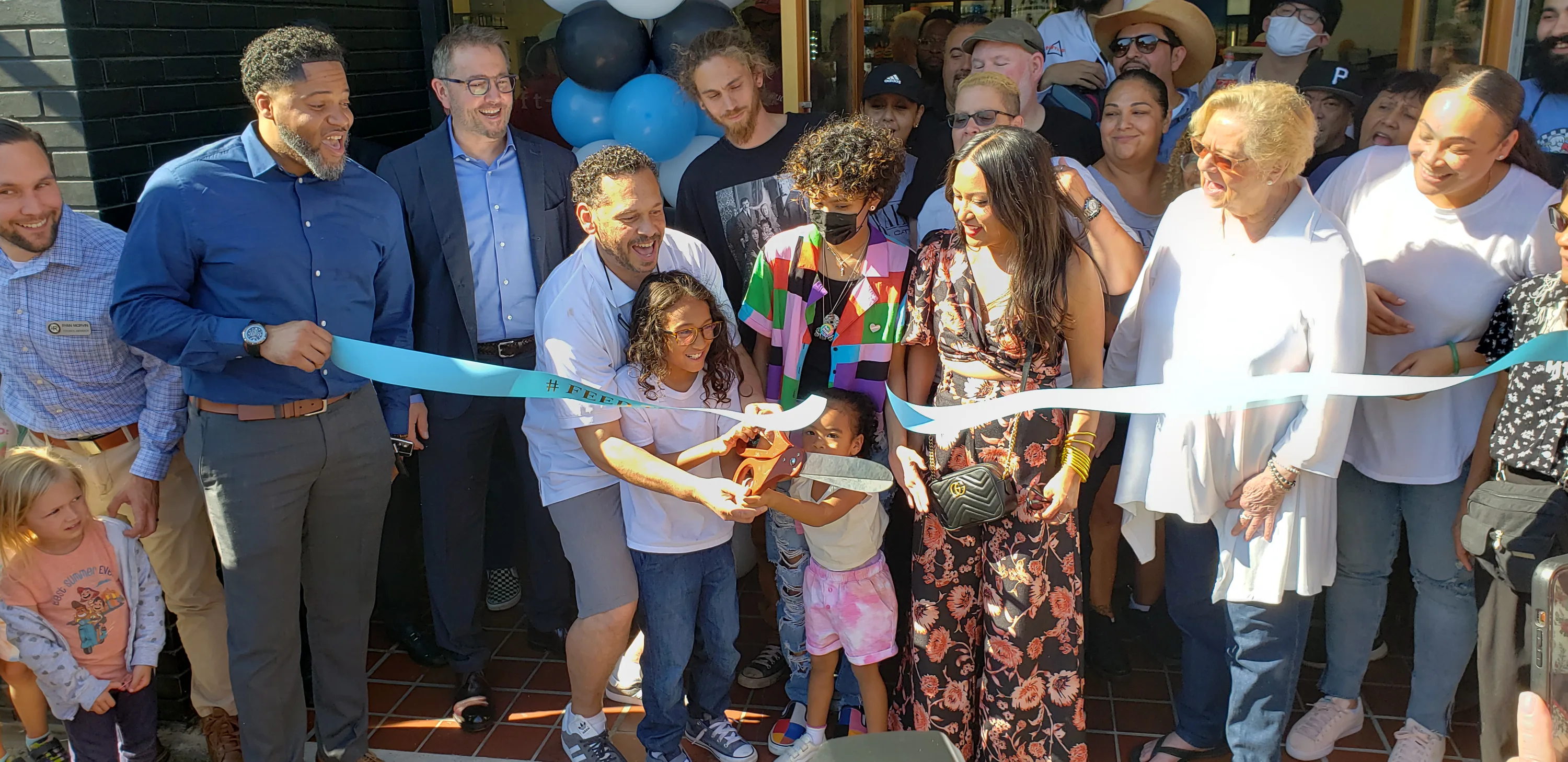 Group of people smiling and gathered for a ribbon-cutting ceremony. A young child helps an adult cut the ribbon with large scissors. Colorful balloons decorate the entrance.