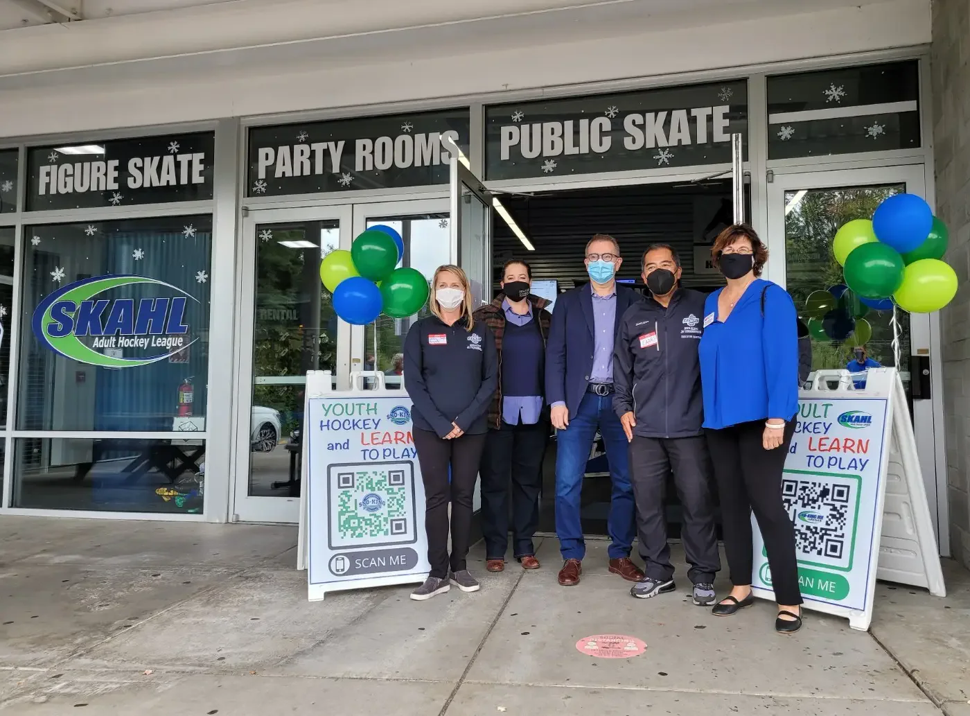 A group of six people wearing masks stand in front of an ice rink entrance with signs for public skating and hockey. Balloons and a QR code for youth hockey are displayed.
