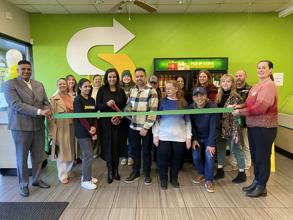 A group of people stand smiling at a Subway sandwich shop's ribbon-cutting event. They hold a green ribbon and large scissors. The background is a vibrant green wall.