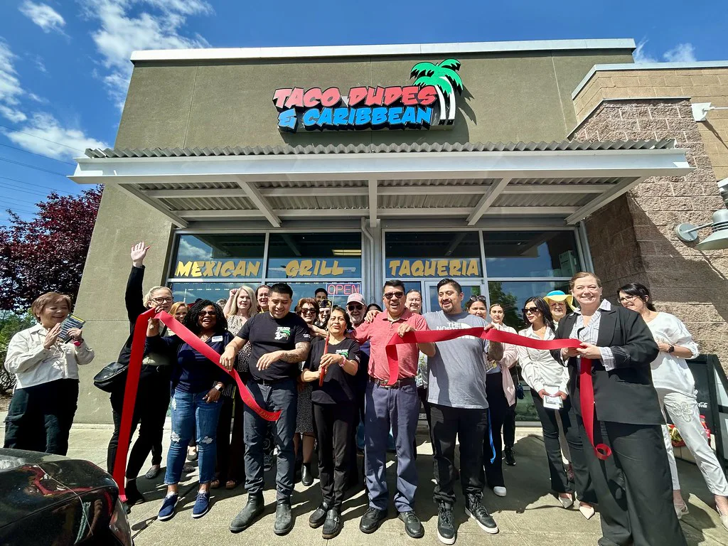 A group of people celebrate the ribbon-cutting ceremony of Taco Dudes & Caribbean, a Mexican Grill and Taqueria, with a red ribbon in front of the restaurant's entrance.