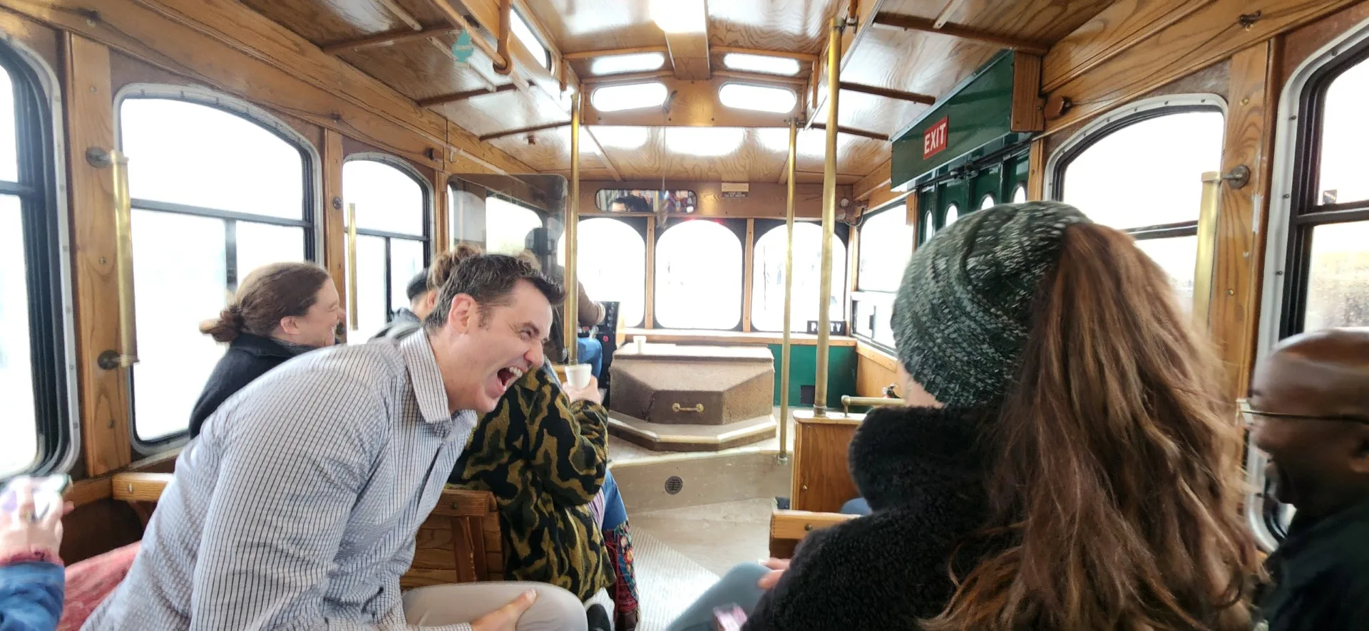 Interior of a wooden trolley with passengers laughing and talking, a man leaning forward mid-laugh