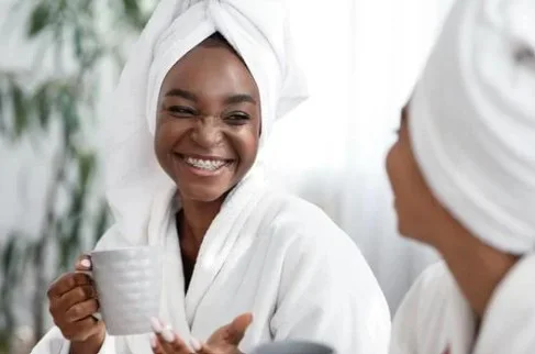 Two women in white bathrobes and hair towels are seated, smiling at each other. One holds a coffee mug, creating a relaxed and cheerful atmosphere.