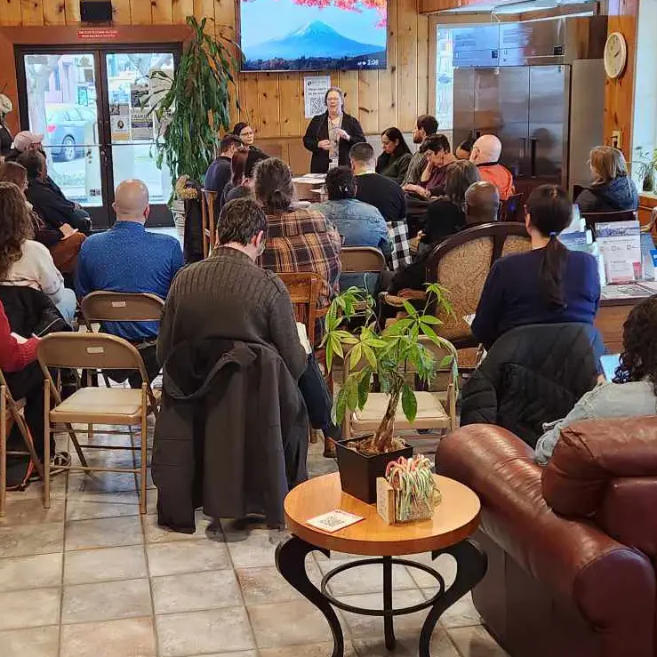A group of people seated in a cozy room with wood-paneled walls, attending a presentation. A woman stands at the front speaking, with posters and brochures visible around.