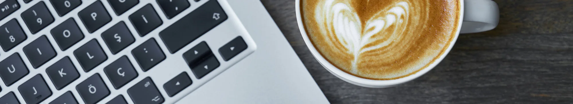 A laptop keyboard next to a cup of cappuccino with heart-shaped latte art, on a dark wooden table.