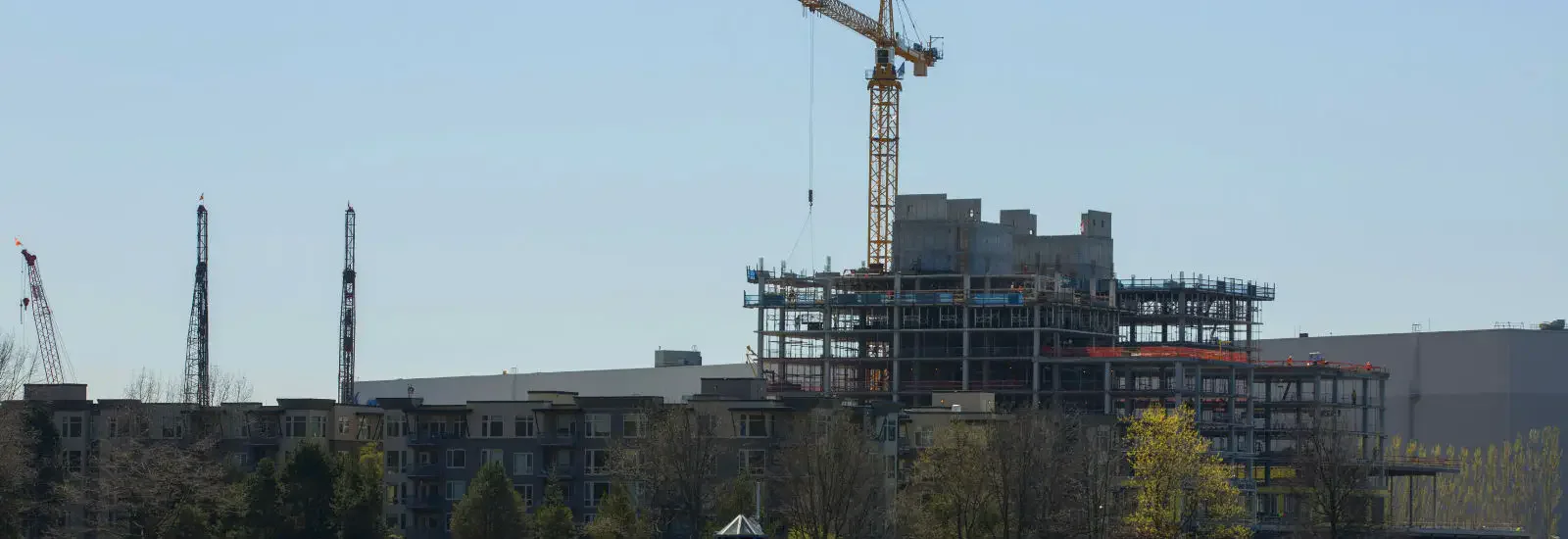 Construction site with a high-rise building under development. A large crane towers above, and apartment buildings are visible in the foreground. Trees line the bottom of the image.