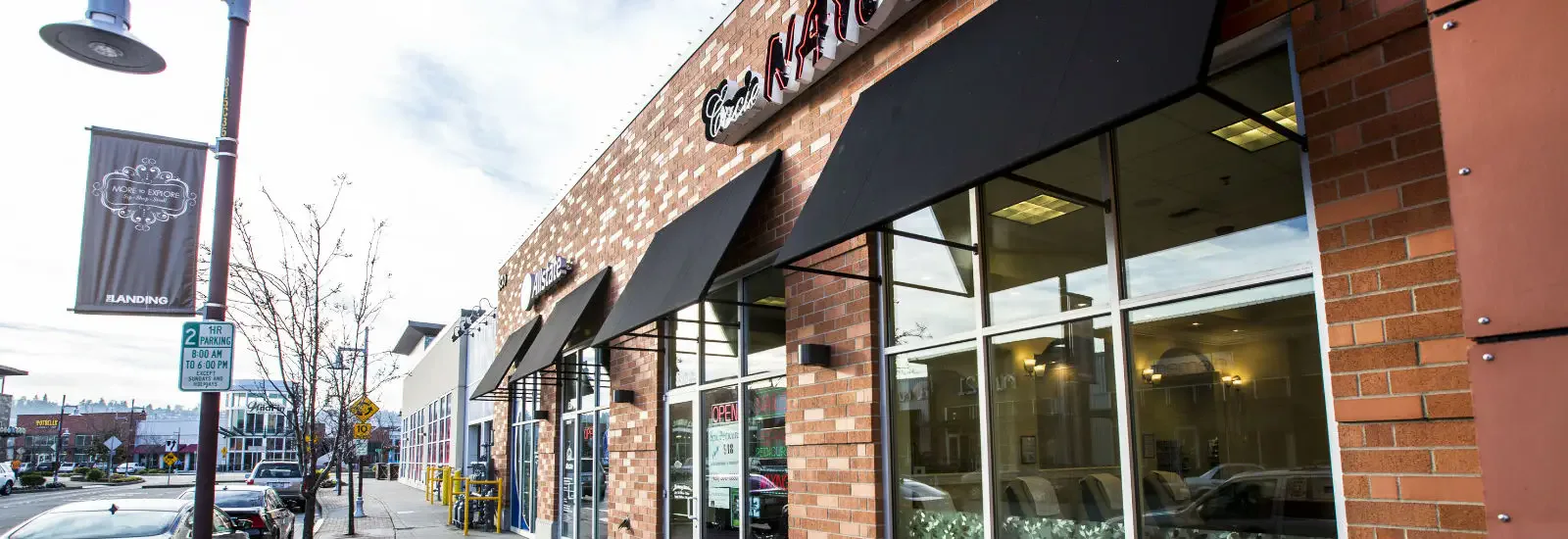 Street view of a commercial area with brick buildings and black awnings. Signs for a nail salon and insurance office are visible. Parking and directional signs are on the sidewalk.
