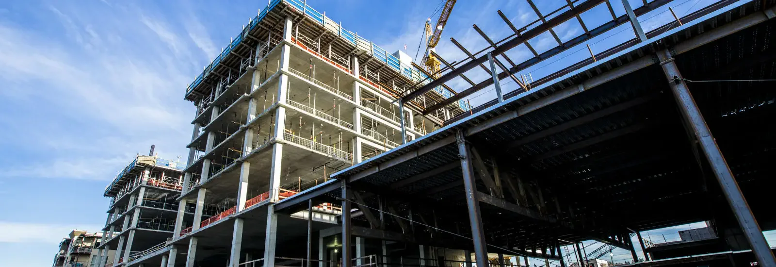 Construction site with a multi-story concrete building framework and a crane under a clear blue sky. Steel beams and scaffolding visible, indicating ongoing development.