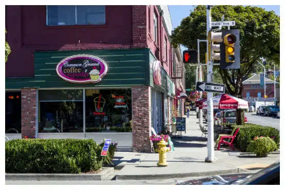 Street corner view with a coffee shop, red brick facade, and neon signage. Traffic lights and a one-way sign are visible. Sidewalk lined with potted plants and seating.