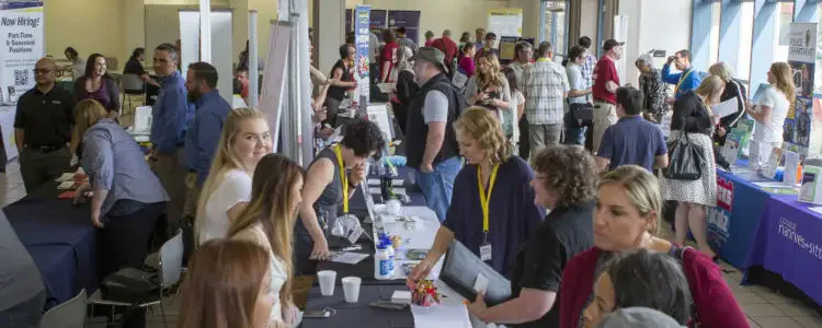 A busy job fair with numerous people visiting various booths. Tables are lined with informational materials, and attendees are engaging with representatives for potential employment.