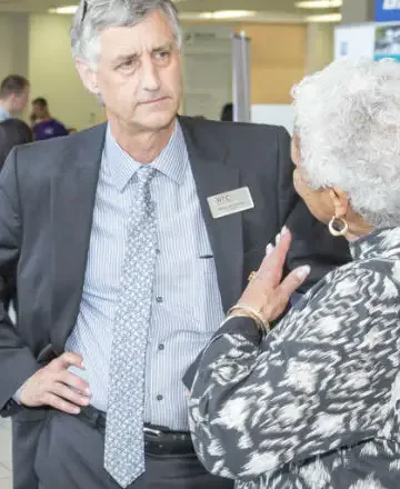 A man in a suit with a nametag and a woman with white hair in a patterned jacket are engaged in a conversation at an indoor event, with others in the background.