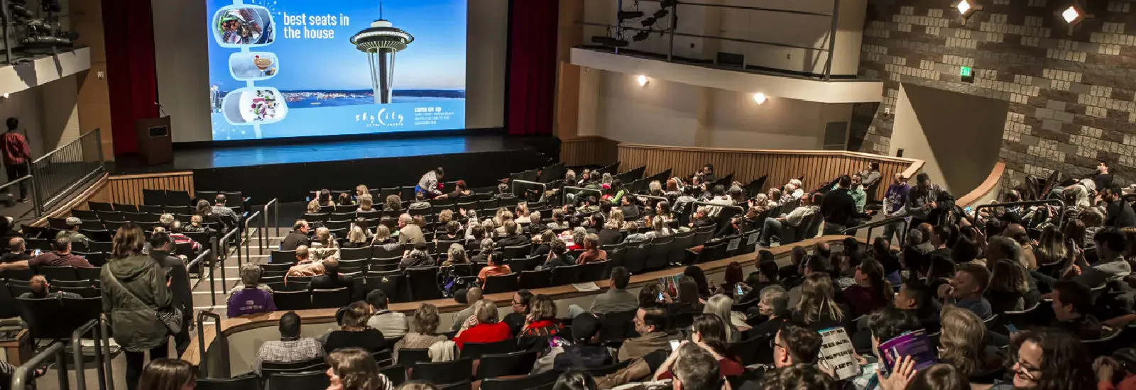 A crowded theater auditorium with people seated, facing a large screen displaying an advertisement featuring the Seattle Space Needle and the text 