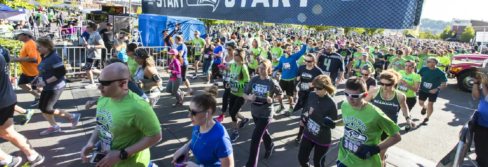 A large group of runners in bright clothing participates in a race, passing under a START banner. The event appears energetic and lively, taking place on a sunny day.