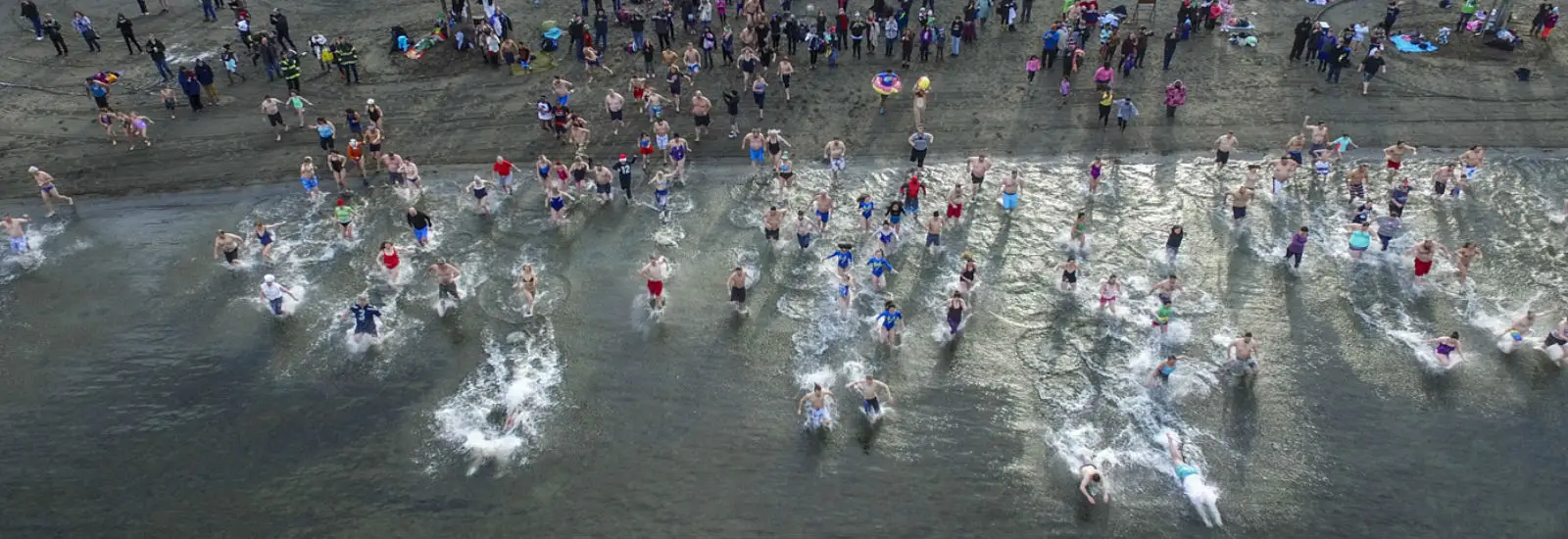 Aerial view of numerous people running into the sea for a winter swim event. Participants are wearing swimsuits, surrounded by spectators on the sandy shore under a cloudy sky.