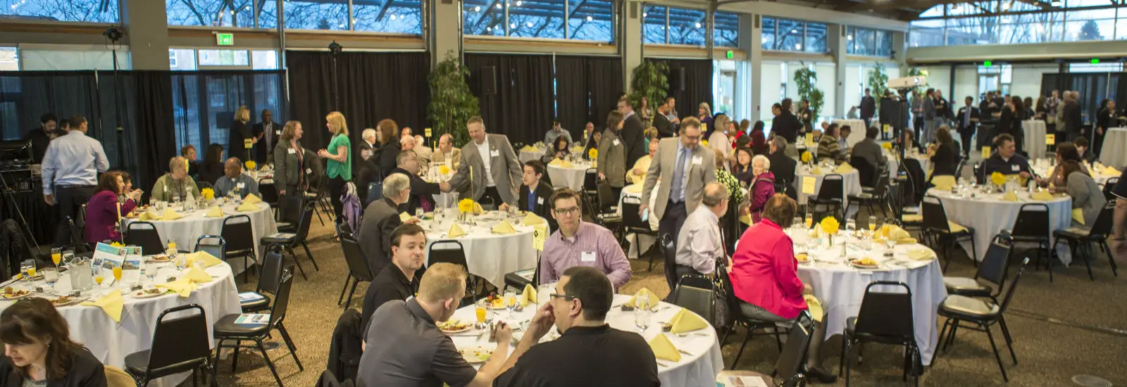 A large banquet hall filled with people seated at round tables, engaged in conversation. Tables are adorned with yellow flowers and napkins. Casual and semi-formal attire.