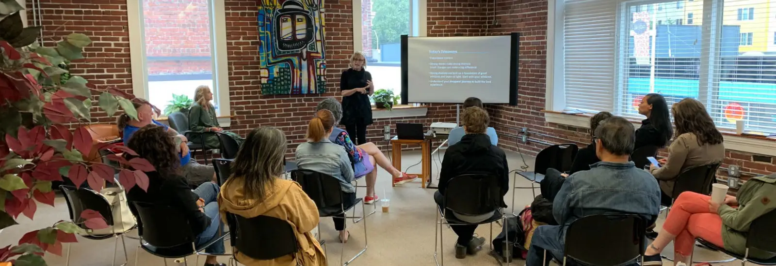 A group of people sit in a room with brick walls, listening to a presentation. The presenter stands near a screen displaying text. Vibrant artwork is on the wall.