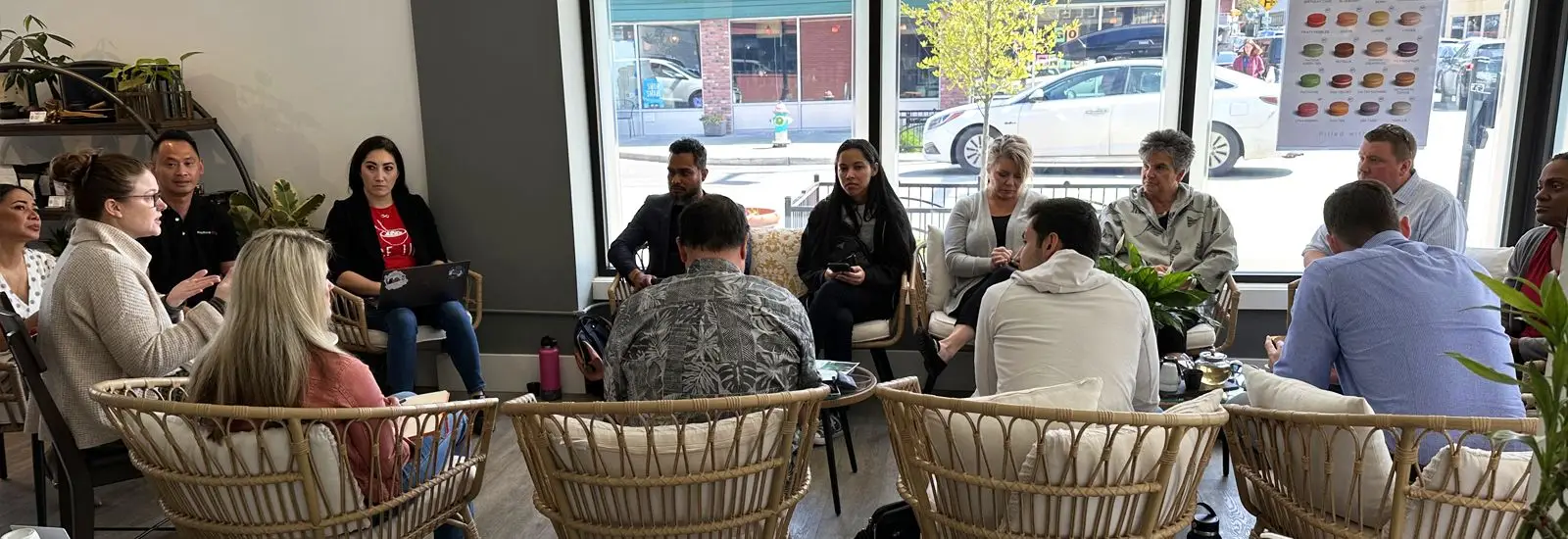 A diverse group of people sits in a circle inside a cafe, engaged in a discussion. Plants and a large window with a street view in the background create a cozy atmosphere.