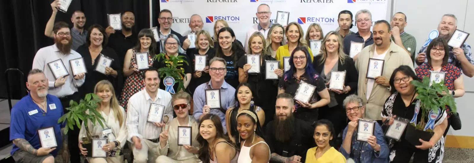 A diverse group of people smiling and holding award plaques and potted plants. They are standing in front of a backdrop with logos for Renton Chamber and Renton Reporter.