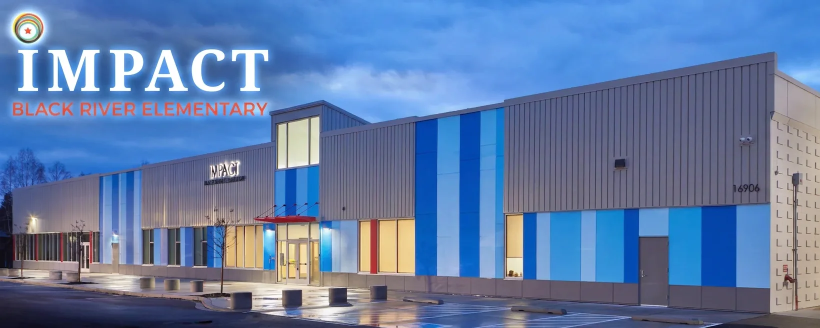 Exterior of IMPACT Black River Elementary school at dusk, modern building with blue vertical panels, illuminated entrance and empty wet parking lot.