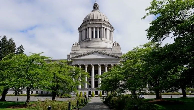 Washington State Capitol building in Olympia with white dome and columns surrounded by green trees on a clear day during the 2026 legislative session.