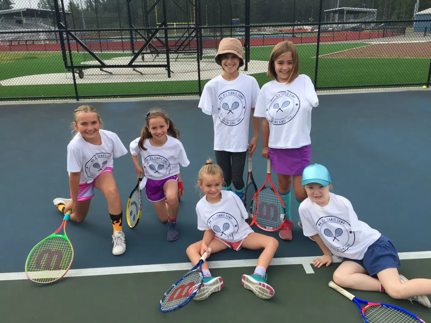 Six children in tennis camp shirts pose on a tennis court, holding rackets and smiling. Four are kneeling, while two stand behind them. A chain-link fence is in the background.