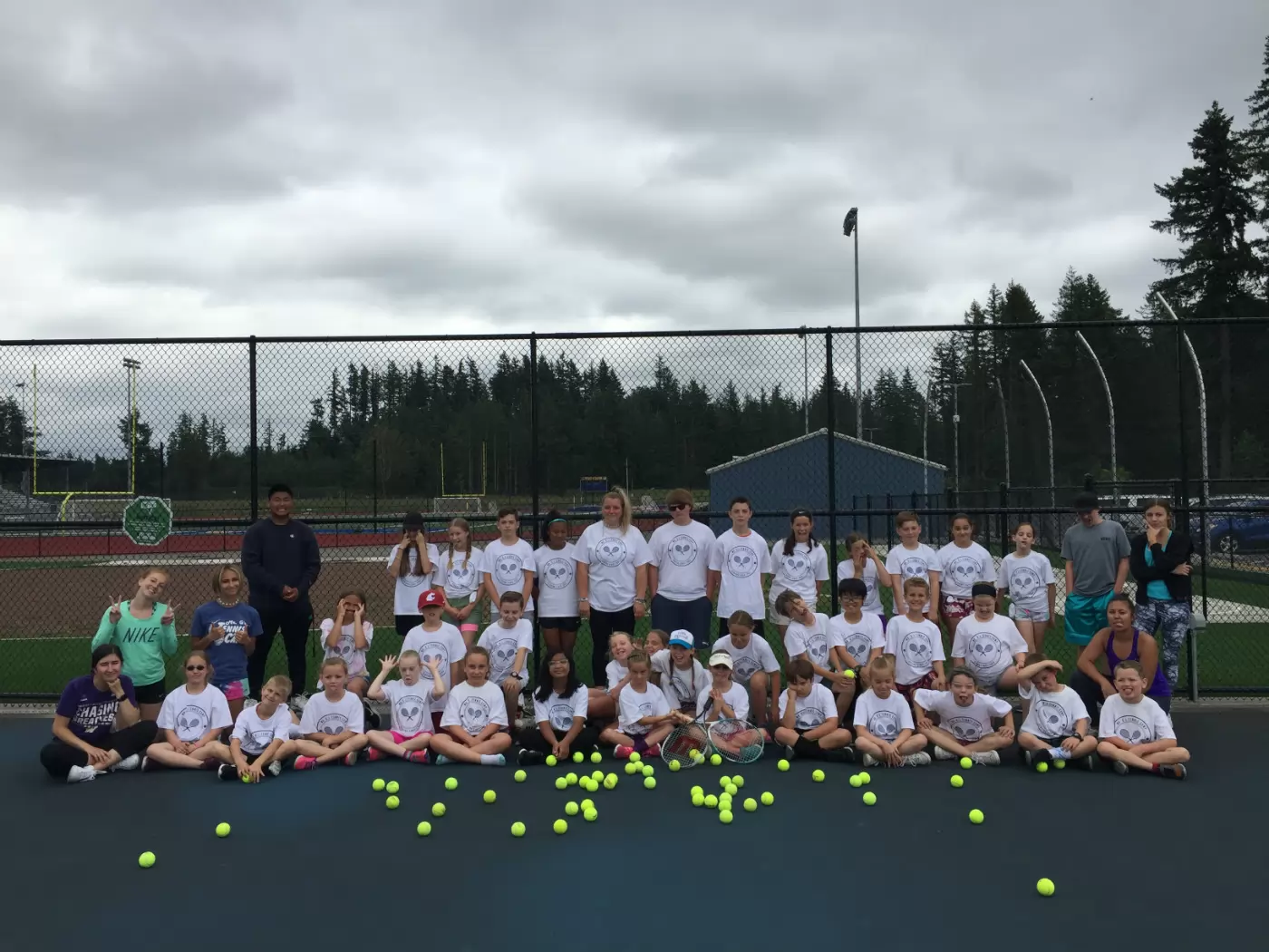 A group of kids and adults in sports attire pose on a tennis court with tennis balls scattered in front. A chain-link fence, trees, and a cloudy sky are in the background.