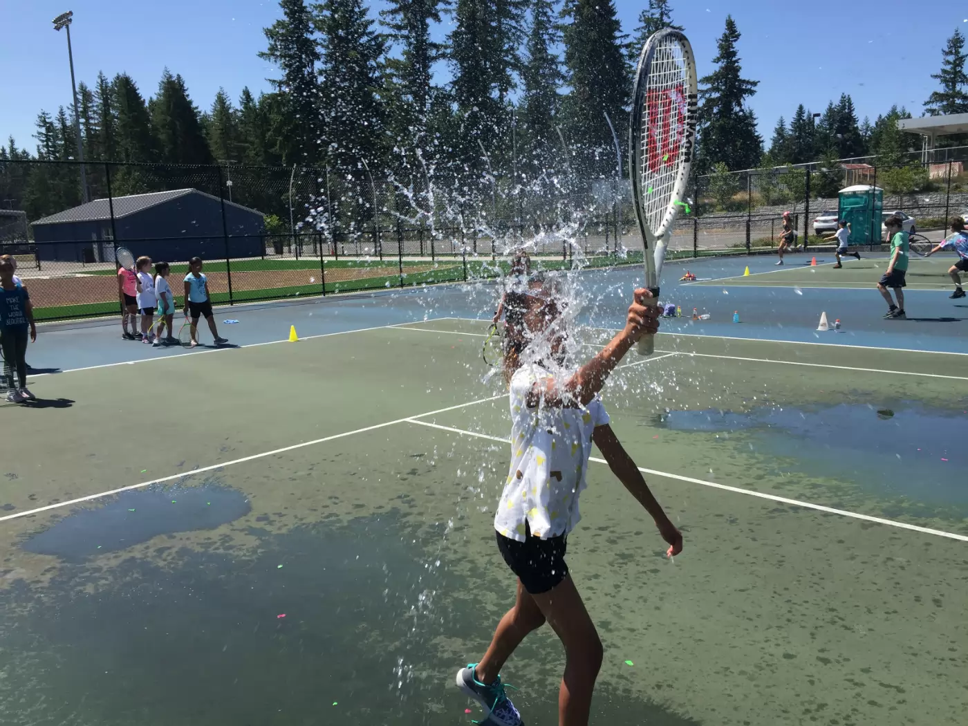 A child swings a tennis racquet, bursting a water balloon on a sunny outdoor court. Other children watch from a distance, with trees and a building in the background.