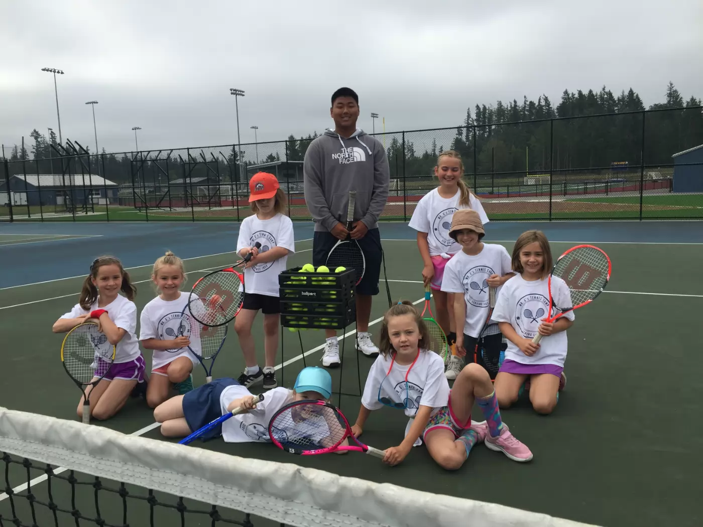 Group of children with tennis rackets and coach pose on a tennis court. They're smiling, wearing sportswear and a variety of hats, with a basket of tennis balls nearby.