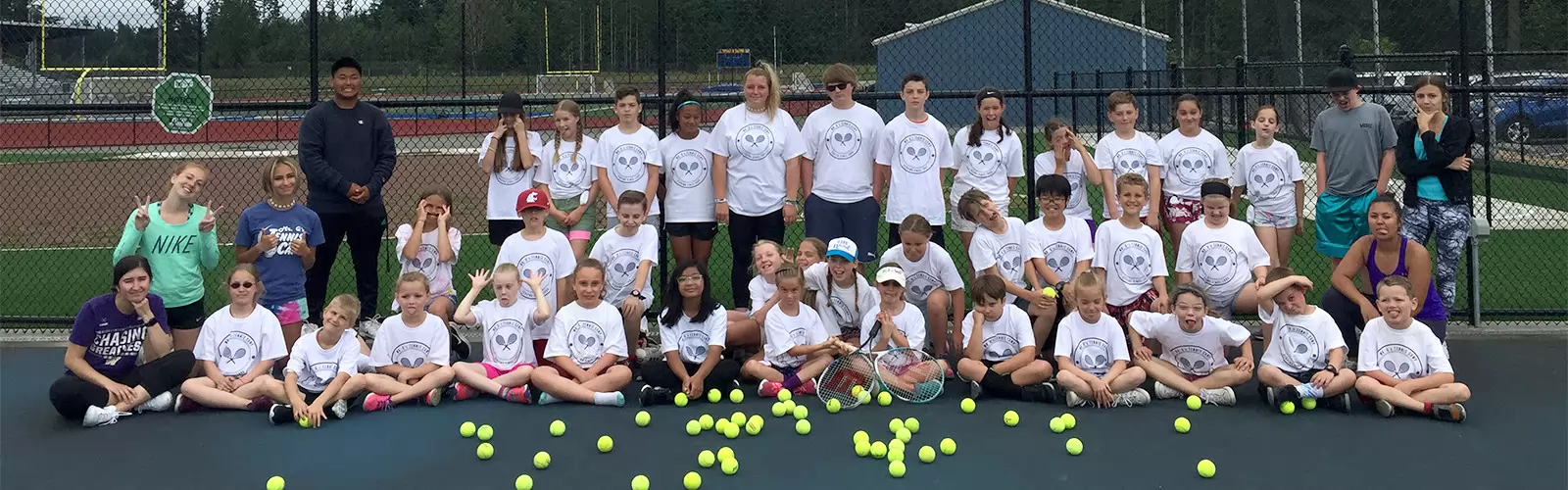 A large group of children and teens wearing matching tennis-themed shirts pose on a tennis court with scattered tennis balls. They are smiling and sitting or standing in rows.