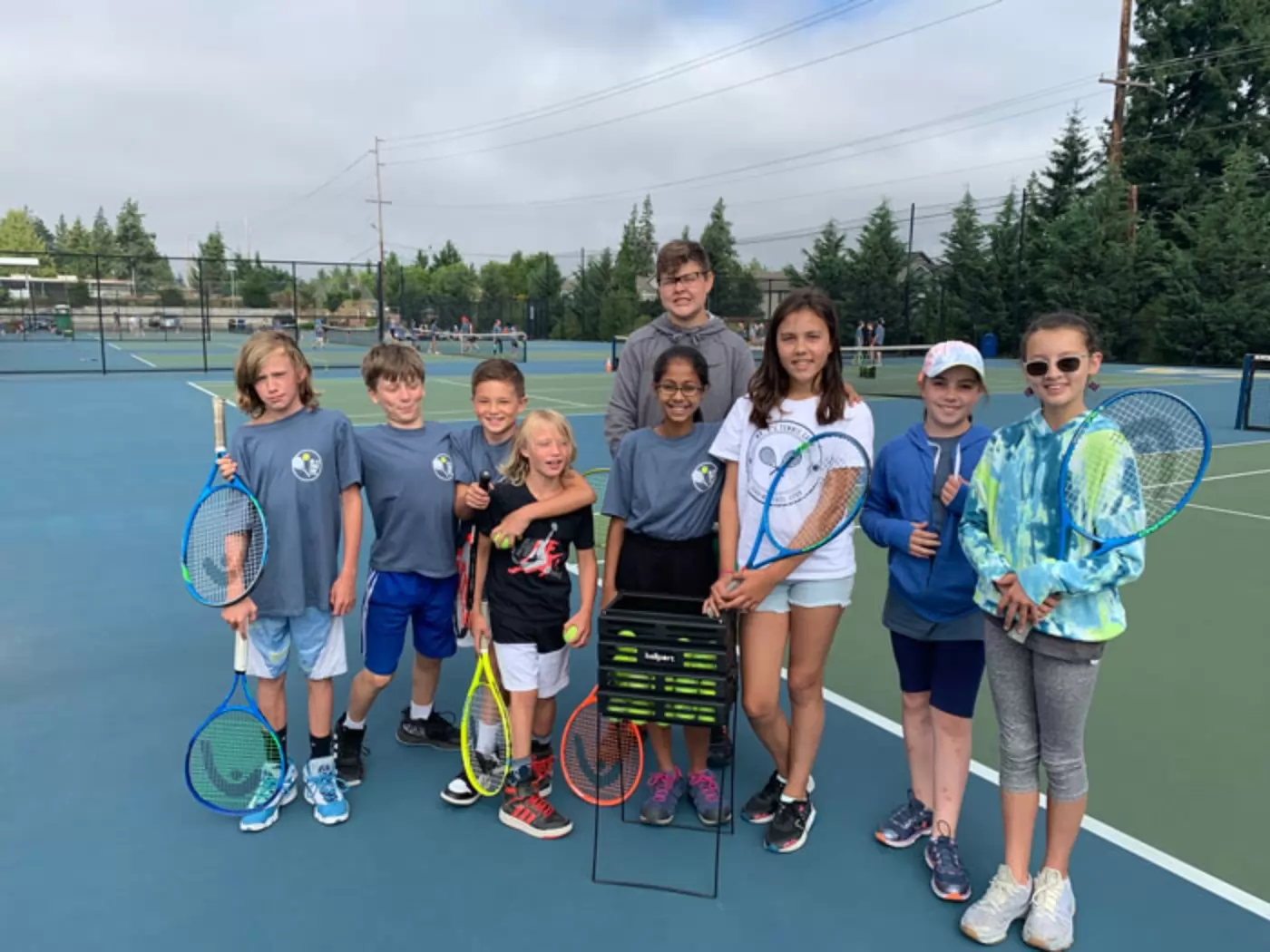 A group of nine children, holding tennis rackets, stands together on a tennis court. They are dressed in casual sportswear and appear to be smiling.