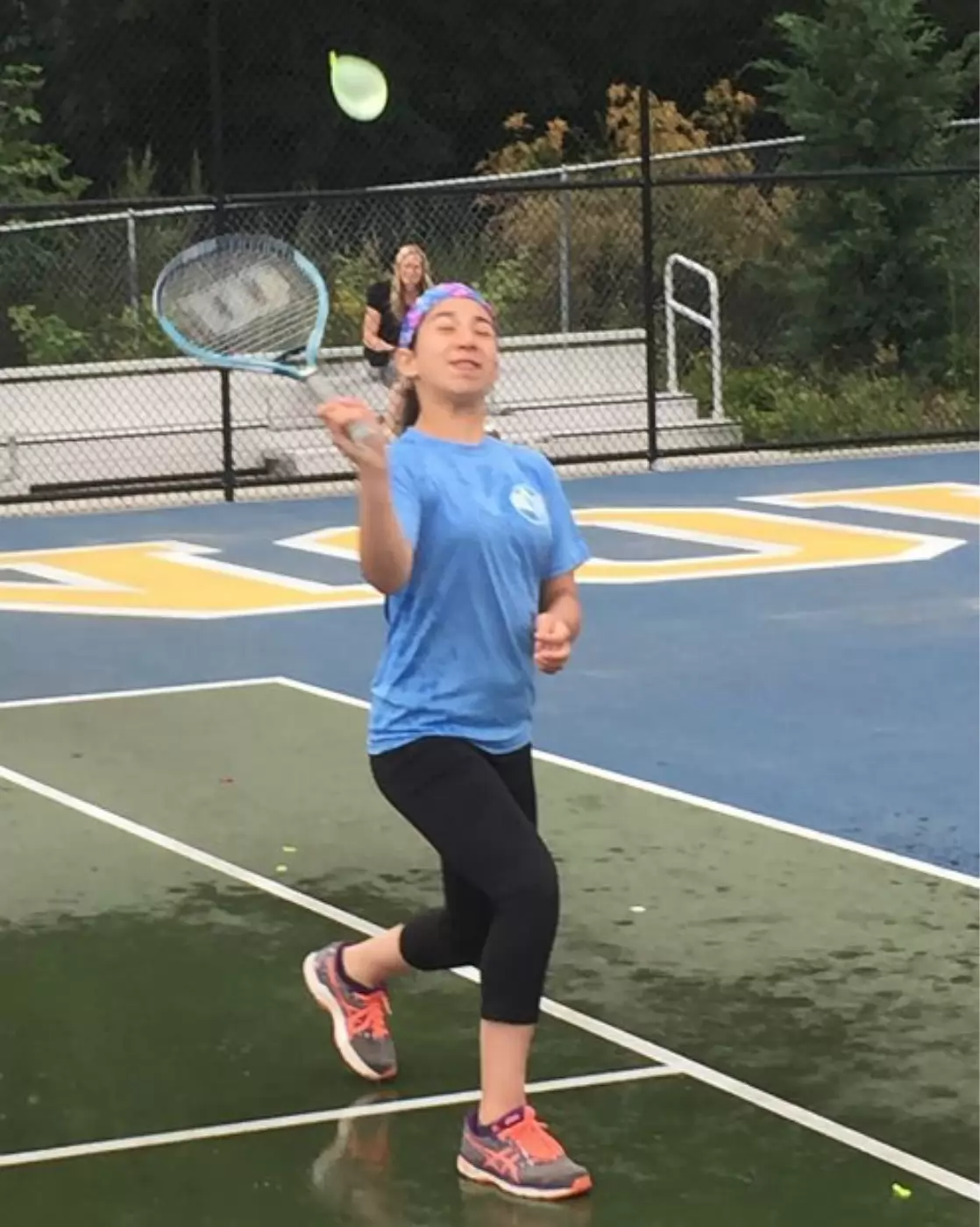 Person in a blue shirt tries to hit a water balloon with a tennis racket on a court, while another person watches in the background. The court appears wet.
