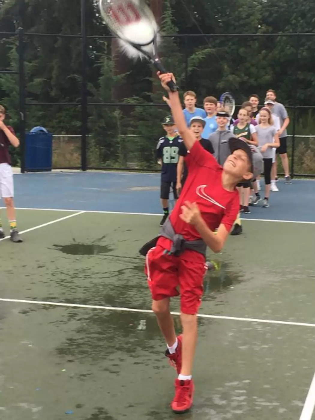 Young boy in red outfit swings a tennis racket at a water balloon on a wet outdoor court. A group of children watch in the background, some holding tennis rackets.
