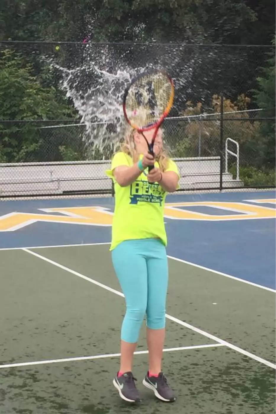 A person in a yellow shirt and blue pants plays tennis, hitting a water-filled balloon with a racket, causing the water to splash dramatically. The scene is on an outdoor court.