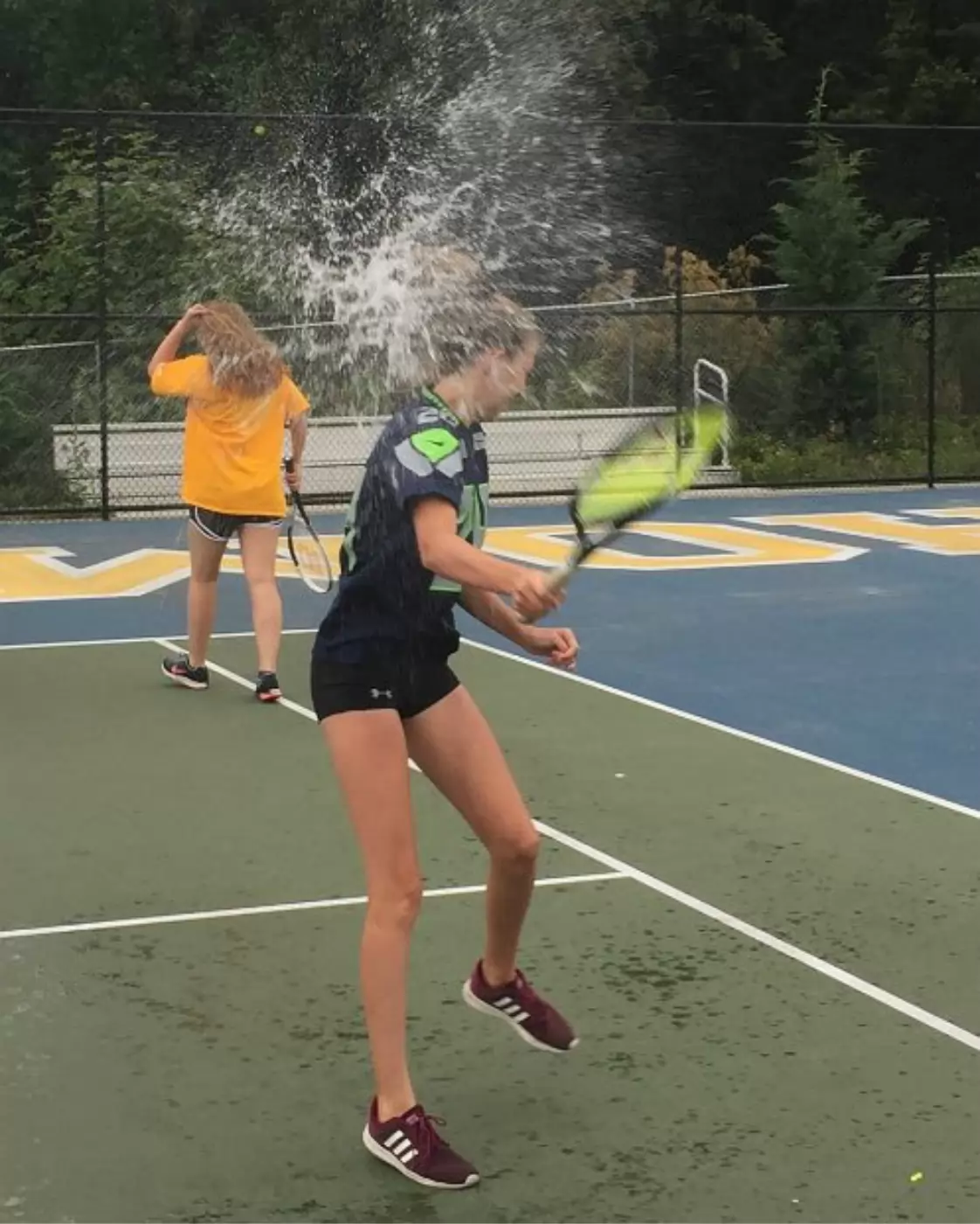 Two girls on a tennis court, one in a blue shirt is getting splashed with water while swinging a racket. The other stands in the background, holding her own racket.