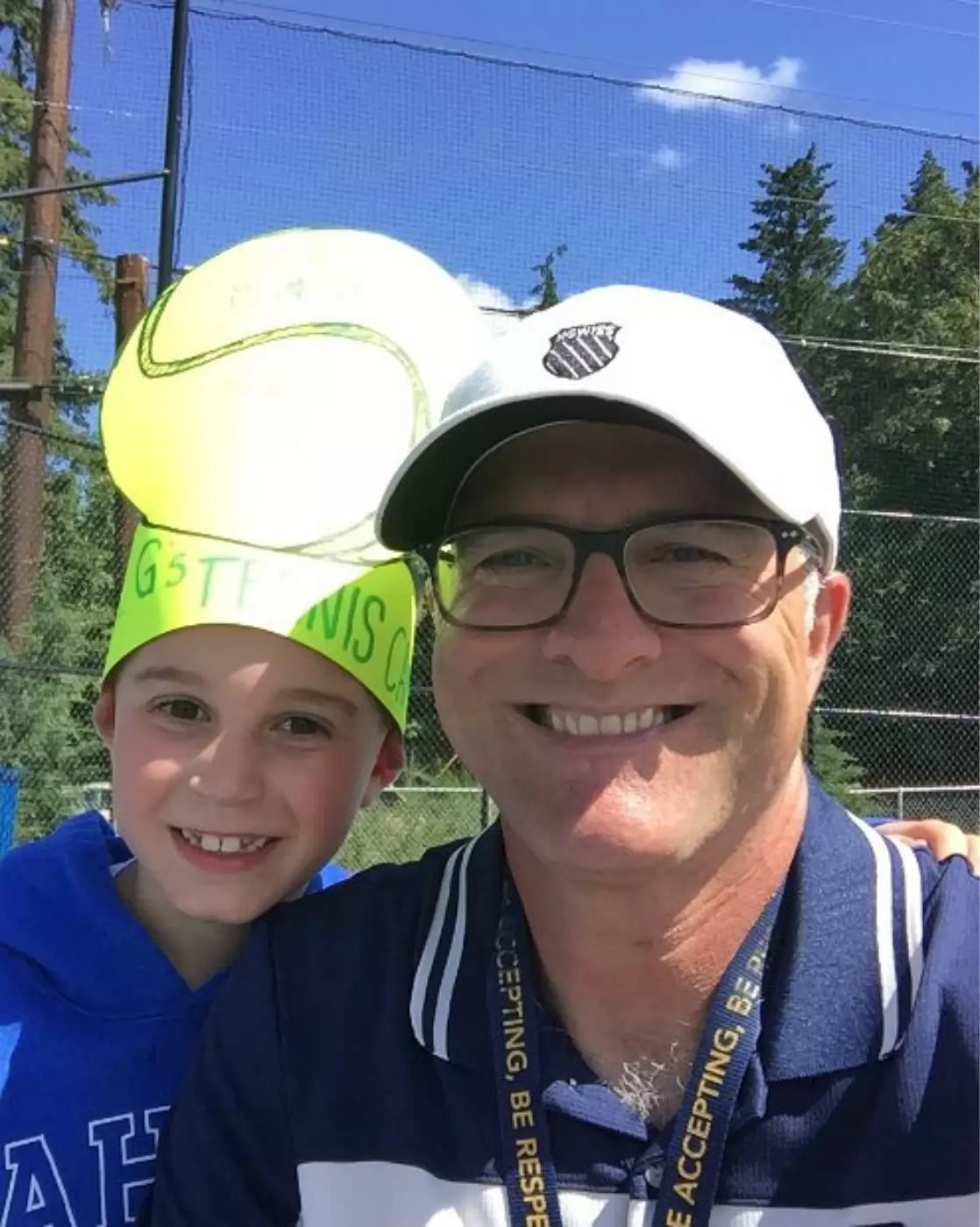 A man and child smiling at the camera outdoors, with the child wearing a large tennis ball-shaped hat. The man is wearing glasses and a white cap. Background has trees and clear sky.
